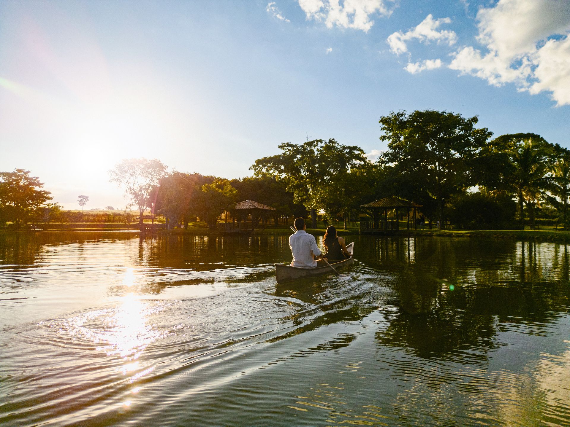 Two people in a rowboat on lake at sunset with trees and a pavilion in the background.