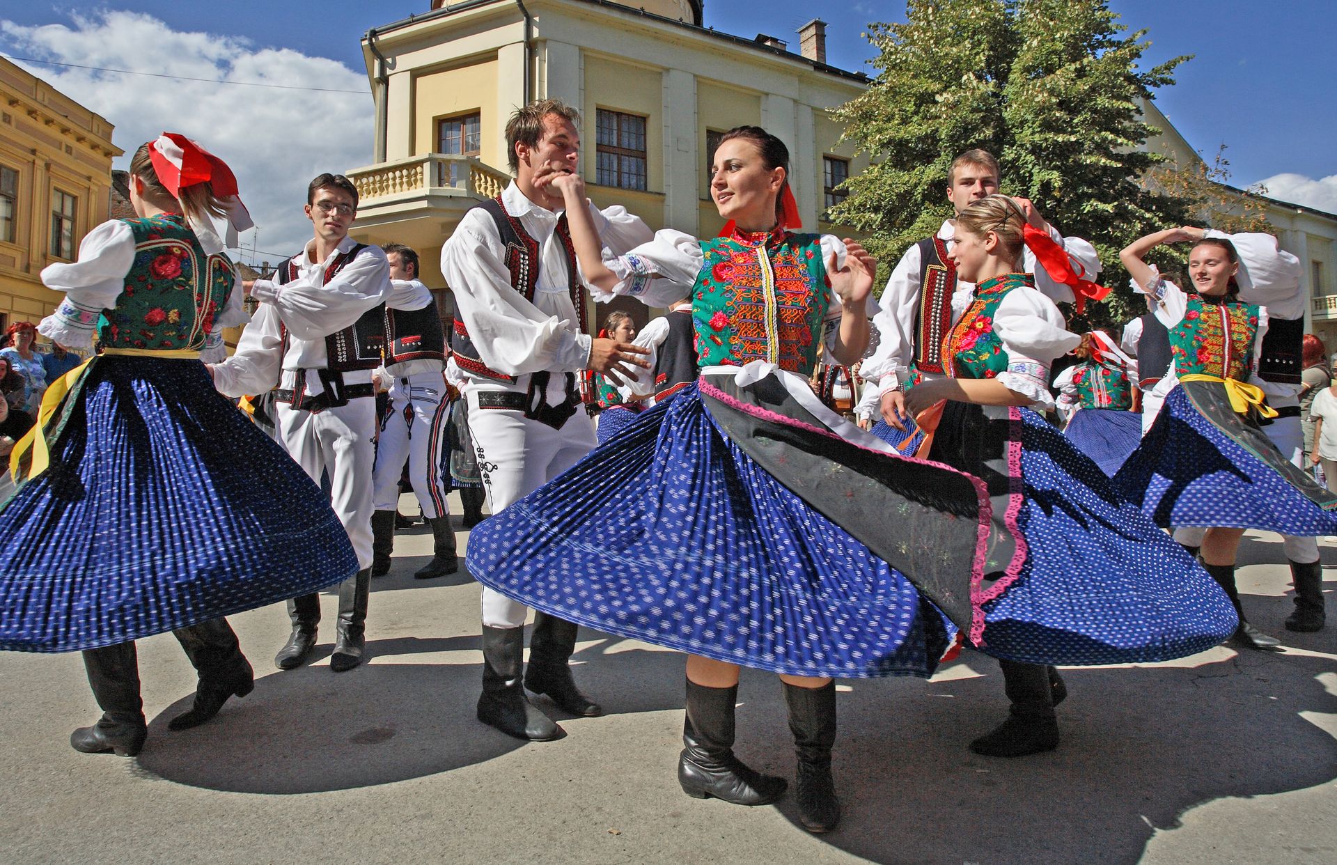 Dancers in colorful traditional Serbian folk costumes perform a lively cultural dance during an outdoor festival, with swirling skirts, embroidered vests, and historic buildings in the background. Dancers in colorful traditional Serbian folk costumes perform a lively cultural dance during an outdoor festival, with swirling skirts, embroidered vests, and historic buildings in the background.
