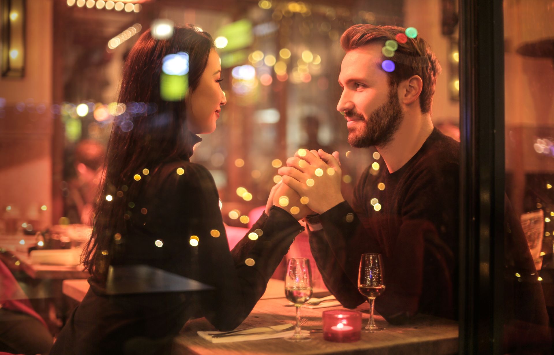 A couple holding hands across a restaurant table, smiling at each other through a softly lit window with warm, romantic lights in the background. A couple holding hands across a restaurant table, smiling at each other through a softly lit window with warm, romantic lights in the background.