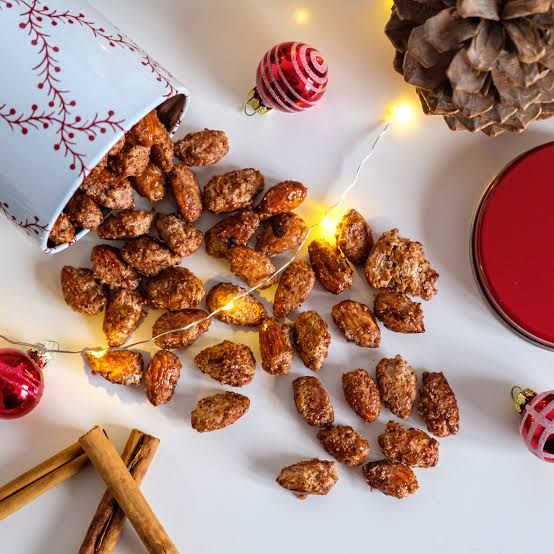 A festive holiday display of freshly roasted German candied almonds spilling from a decorative Christmas mug, surrounded by cinnamon sticks, ornaments, pinecones, and warm string lights. The sweet, cinnamon-coated almonds are a signature treat found at Germany's enchanting Christmas markets and are a must-try during a Christmas river cruise. A festive holiday display of freshly roasted German candied almonds spilling from a decorative Christmas mug, surrounded by cinnamon sticks, ornaments, pinecones, and warm string lights. The sweet, cinnamon-coated almonds are a signature treat found at Germany's enchanting Christmas markets and are a must-try during a Christmas river cruise.
