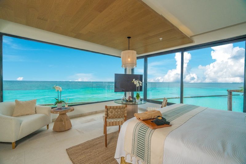 Luxury overwater bungalow interior at Palafitos with king bed, modern furnishings, floor-to-ceiling windows, and panoramic views of turquoise Caribbean waters