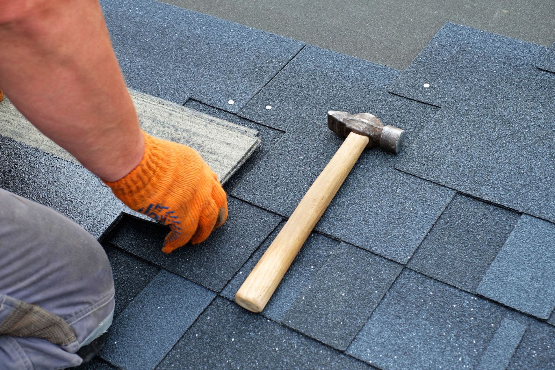 Roofer in Ogden installing asphalt shingles with a hammer, wearing an orange glove during a residential roofing project. Roofer in Ogden installing asphalt shingles with a hammer, wearing an orange glove during a residential roofing project.