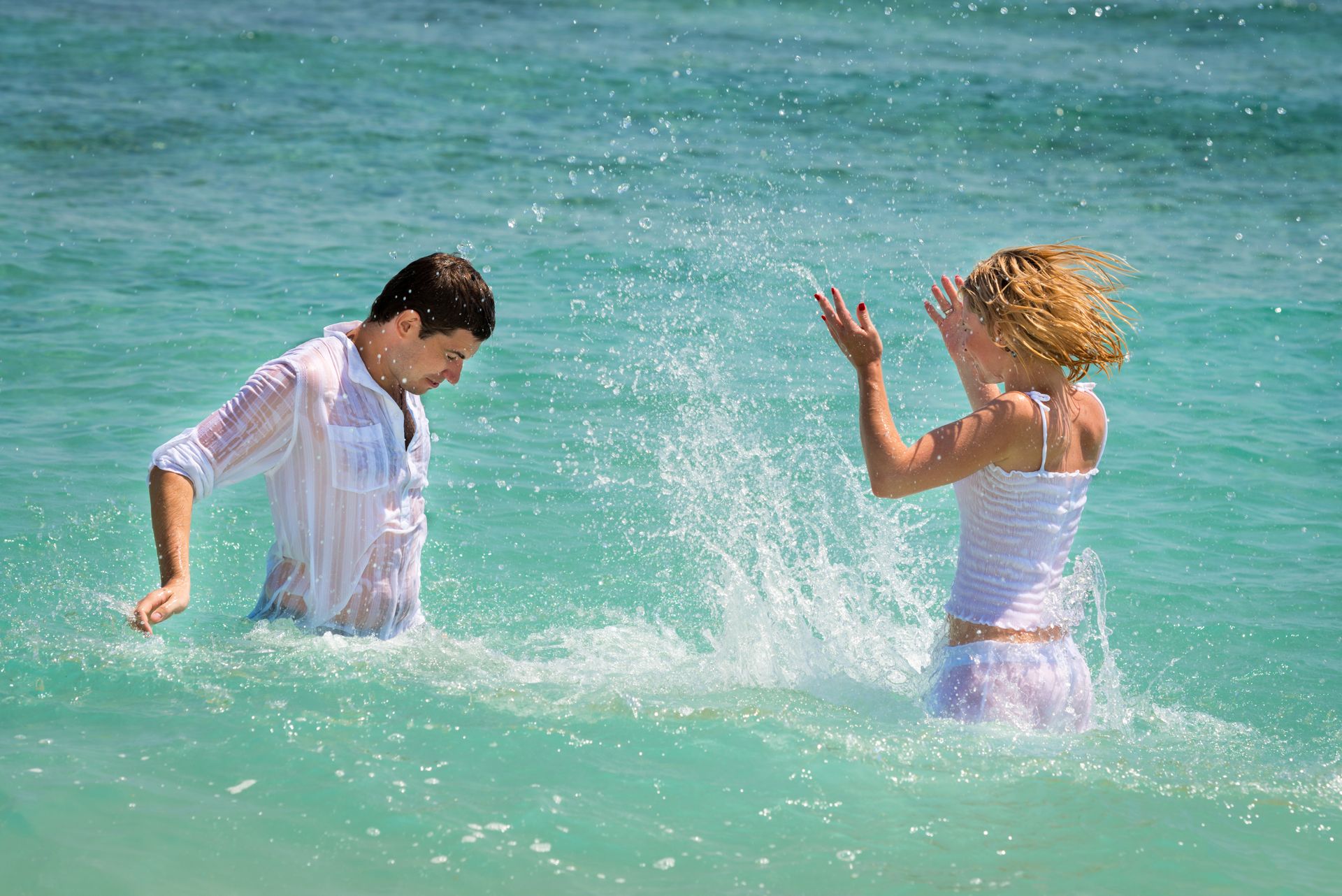 A couple splashes and laughs together in clear turquoise ocean water, wearing white summer outfits and enjoying a sunny tropical day. A couple splashes and laughs together in clear turquoise ocean water, wearing white summer outfits and enjoying a sunny tropical day.
