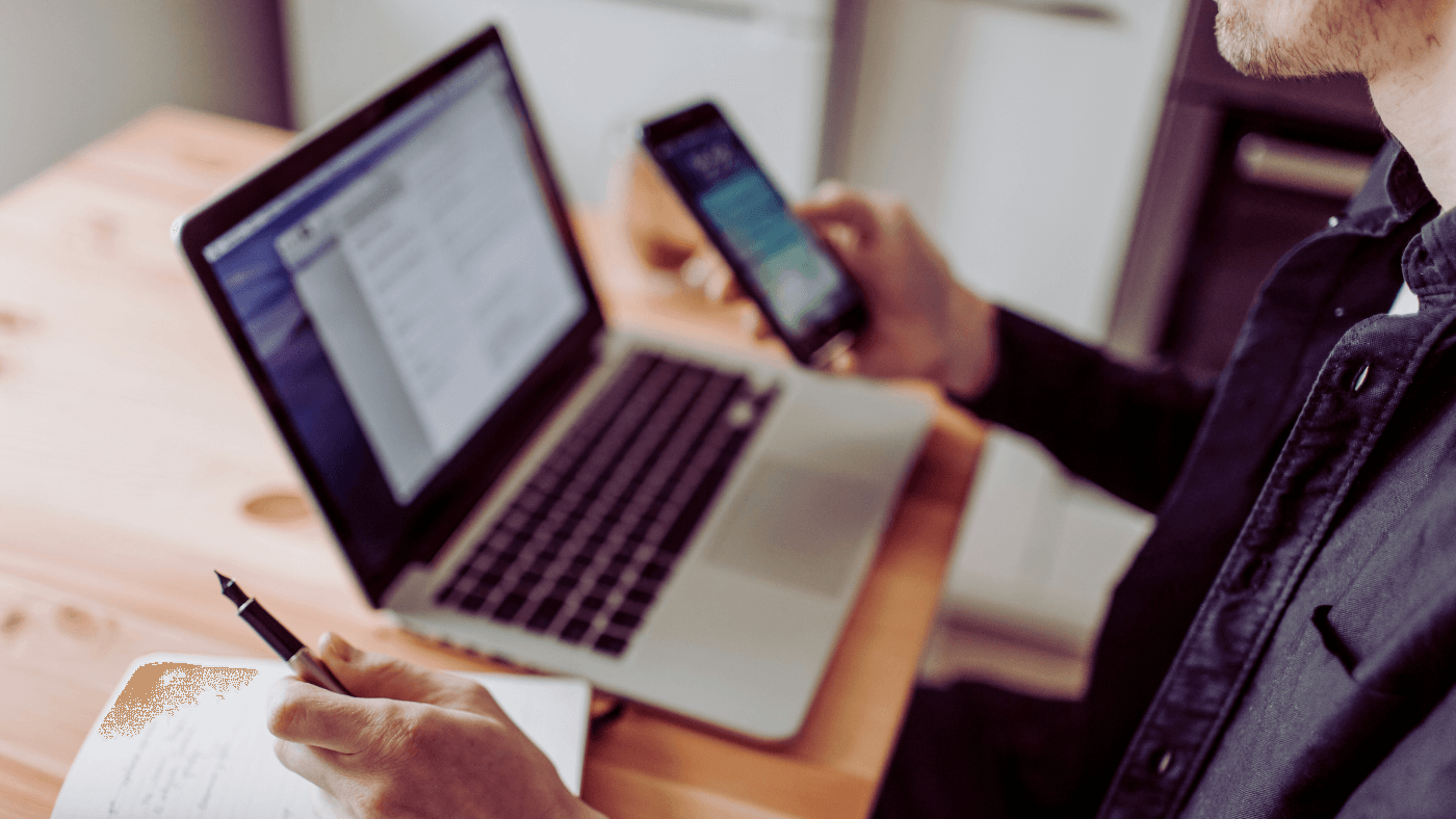 Homem de blusa social com uma caneta na mão esquerda e celular na mão direta. Sobre a mesa, um laptop