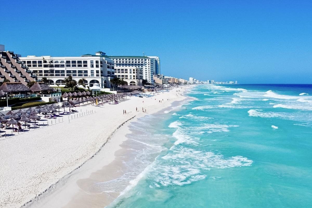 Wide sandy beach in Costa Maya with turquoise waves rolling in, rows of palapa umbrellas and lounge chairs, and beachfront resort buildings lining the shore under a clear blue sky.