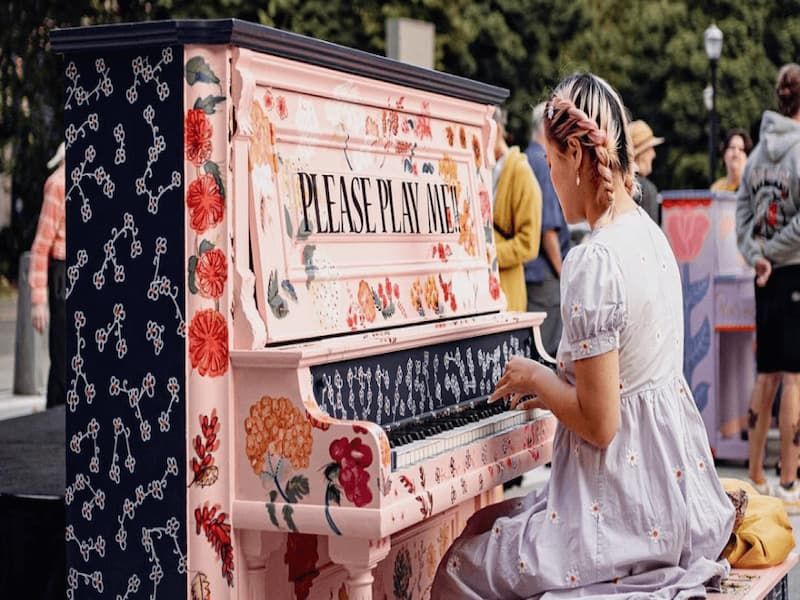 person with braided hair and white daisy dress plays upright piano painted pink and black and covered in painted flowers plus the words 
