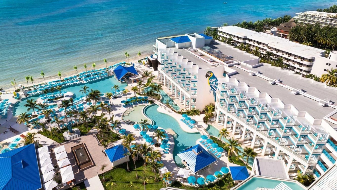 Aerial view of a beachfront resort with turquoise pools, palm trees, and blue-roofed cabanas along the ocean.