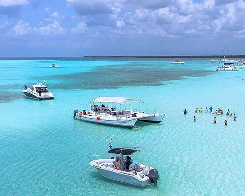 Boats anchored in crystal-clear turquoise water in Cozumel, with people wading and snorkeling in a shallow lagoon under a bright, partly cloudy sky.