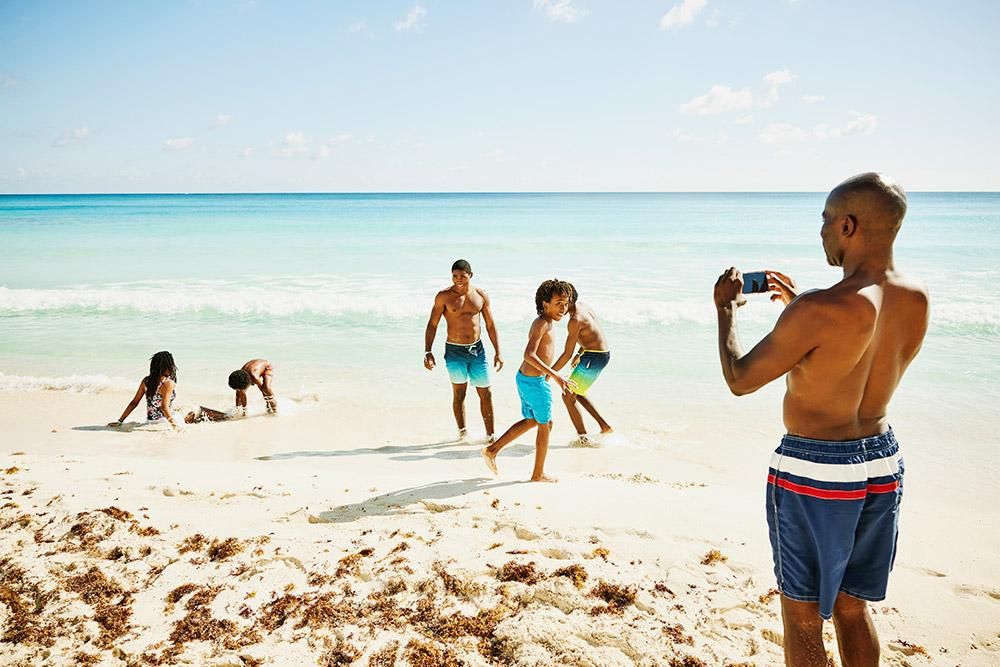 Father photographing children playing on white sand Caribbean beach with turquoise water, kids building sandcastles and running along the shore during family vacation