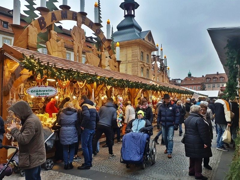 Visitors stroll through the festive Bamberg Christmas Market in Germany, browsing glowing wooden stalls decorated with holiday greenery and traditional Christmas lights.