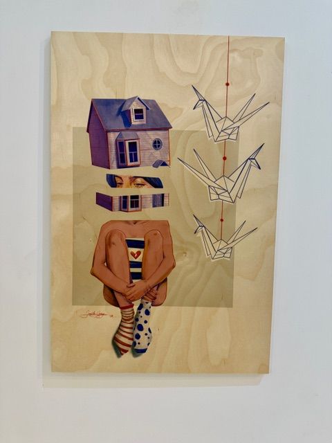 Surrealist mixed media on wood by Jane the Stranger — seated figure with Victorian house as head, origami cranes on red thread beside, wood grain background