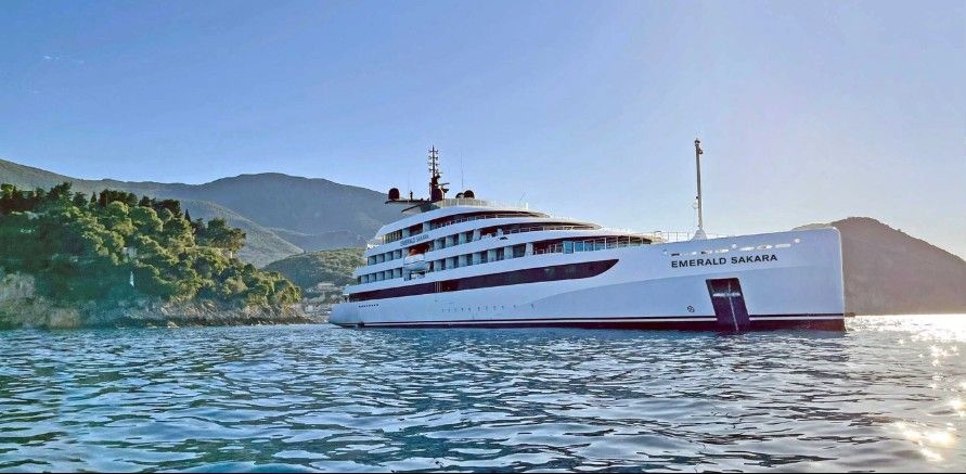 The Emerald Sakara luxury yacht sails on calm blue waters near a rugged, green coastline with mountains rising in the background under a clear sky. The Emerald Sakara luxury yacht sails on calm blue waters near a rugged, green coastline with mountains rising in the background under a clear sky.