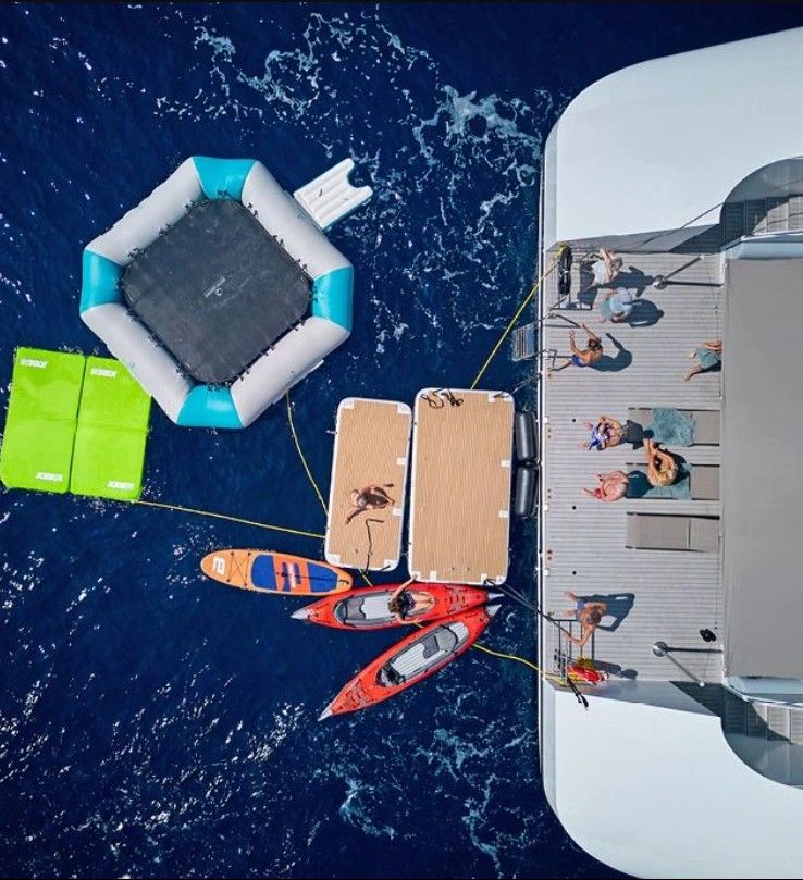 Aerial view of guests enjoying the marina platform from a luxury yacht, with kayaks, paddleboards, a floating trampoline, and deep blue ocean waters surrounding the vessel. Aerial view of guests enjoying the marina platform from a luxury yacht, with kayaks, paddleboards, a floating trampoline, and deep blue ocean waters surrounding the vessel.