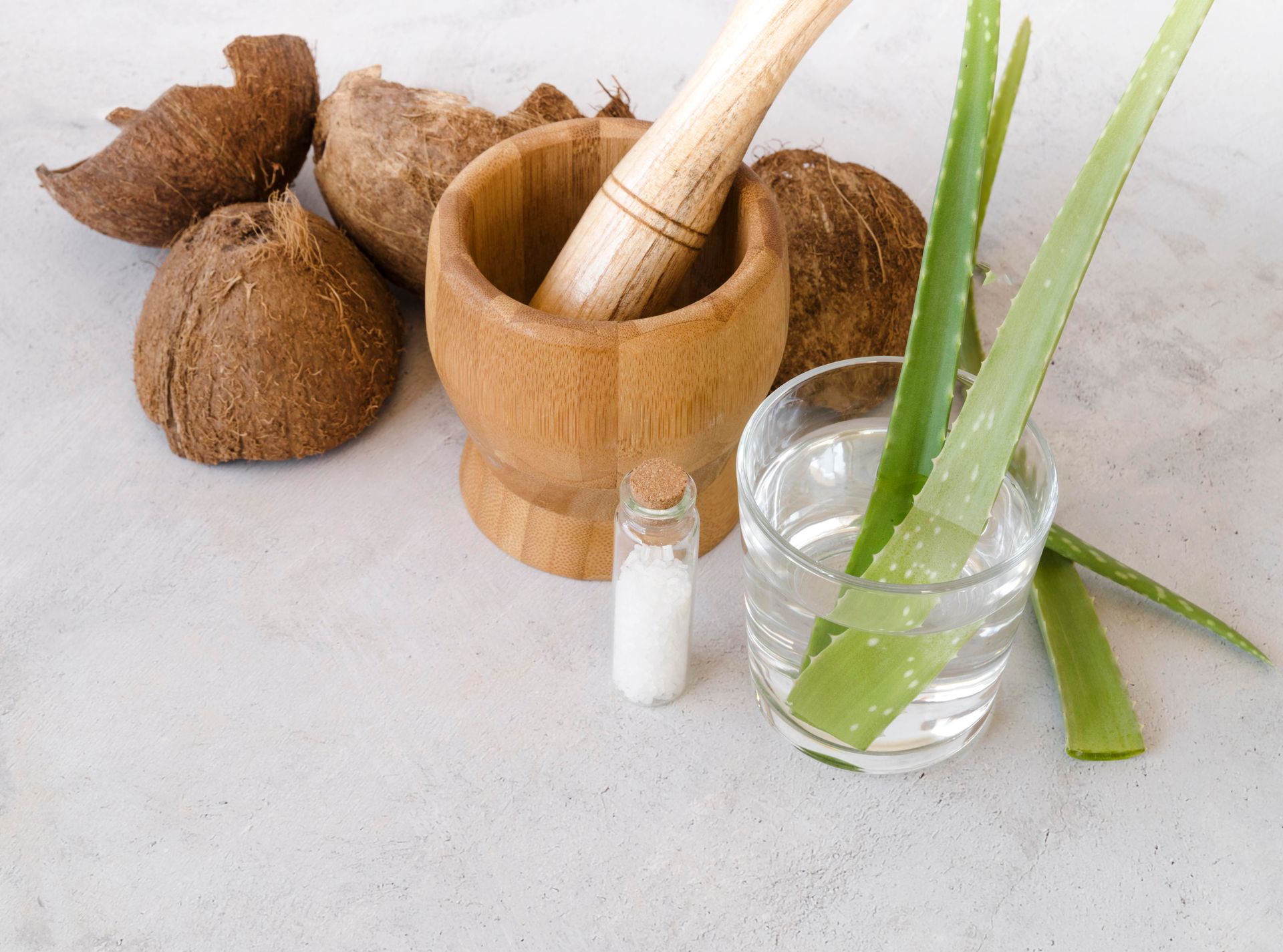 Des branches d'Aloe Vera dans un verre d'eau, une fiole avec un produit blanc dedans, des noix de coco et un mortier pour moudre la coco, poser sur un plan de travail gris béton