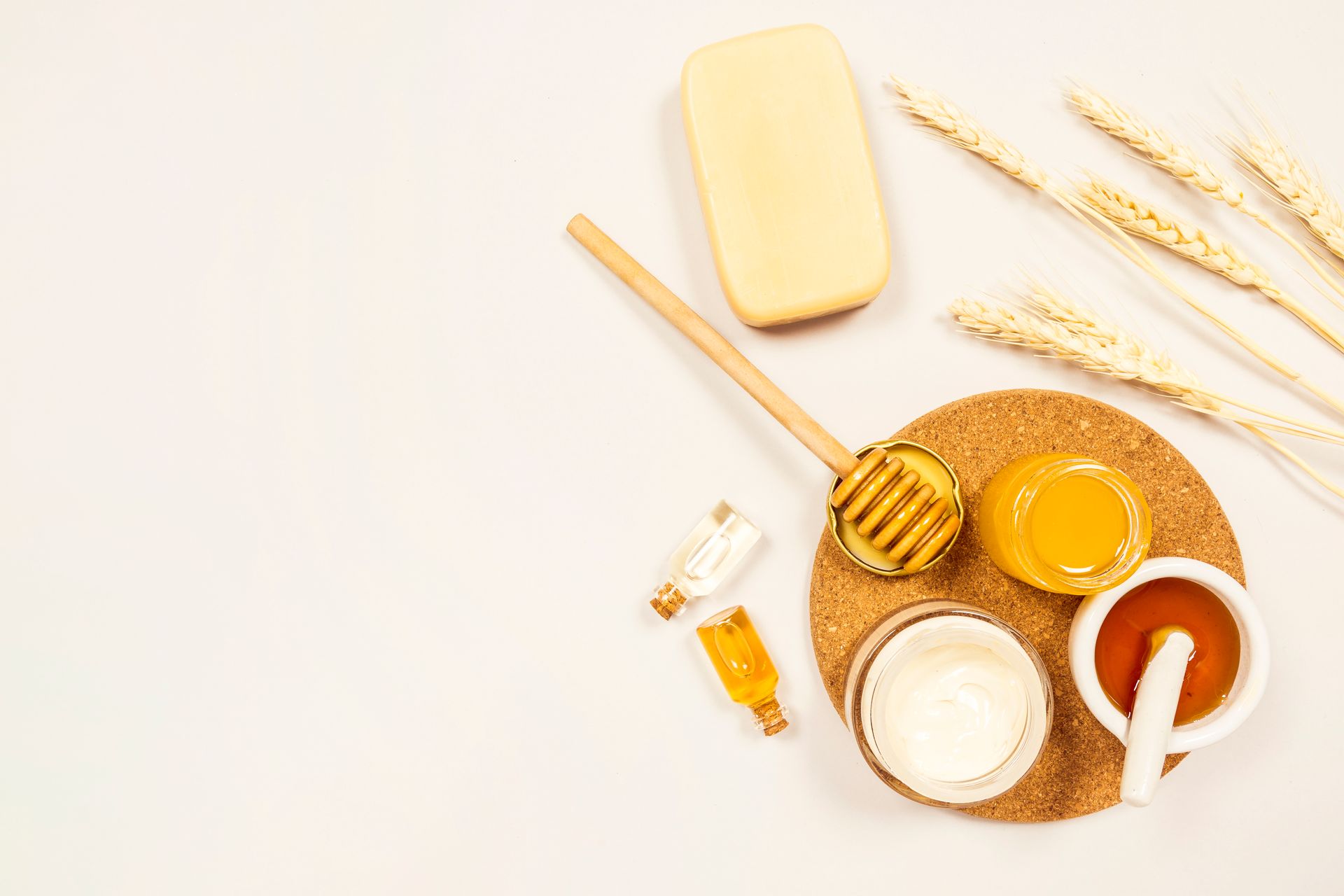 pot de miel doré avec cuillère en bois, flacons d'huiles essentielles, yaourt blanc, savon jaune, épis de blé et sous-verre en liège sur fond blanc lumineux.