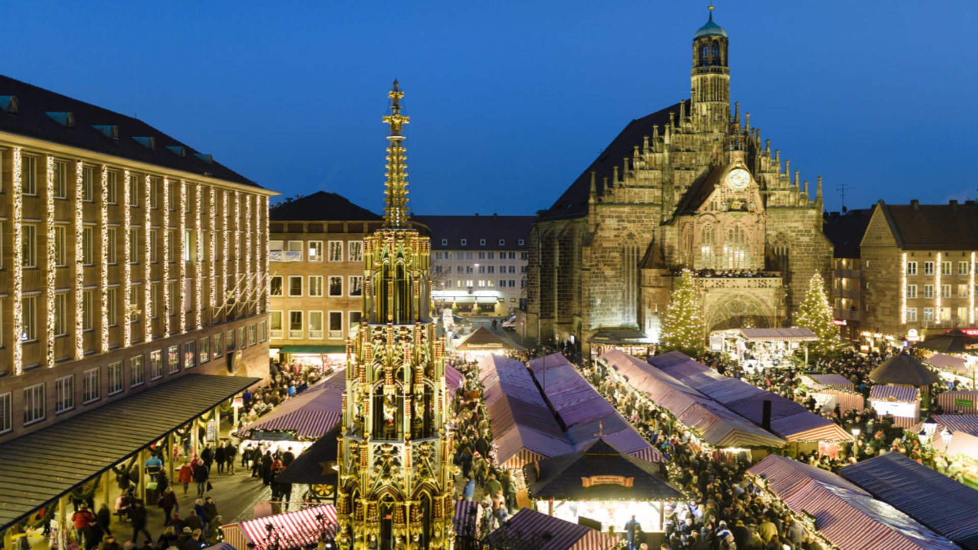 Crowds explore the illuminated Nuremberg Christmas Market at night, surrounded by festive wooden stalls, glowing holiday lights, and historic cathedral architecture in Germany.