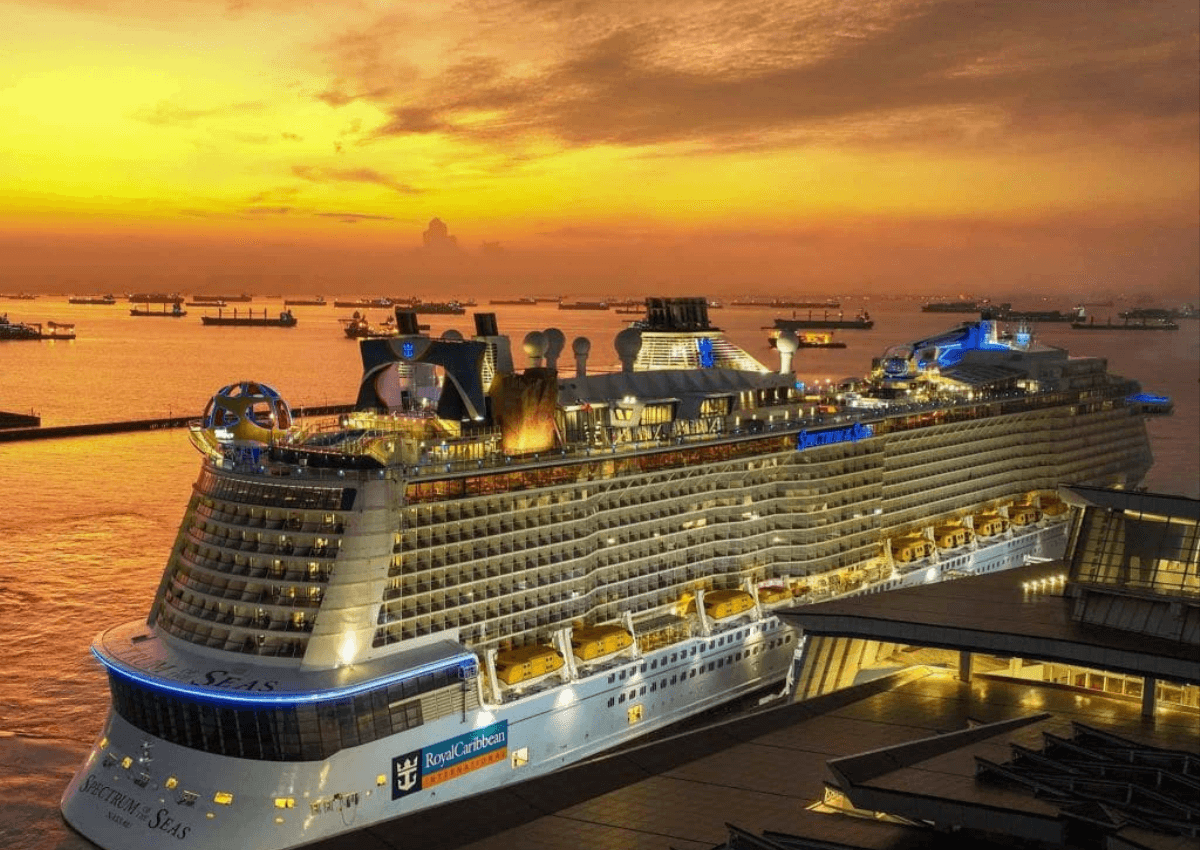 A large cruise ship docked at sunset with glowing lights, calm water, and a golden sky filled with anchored boats in the distance.