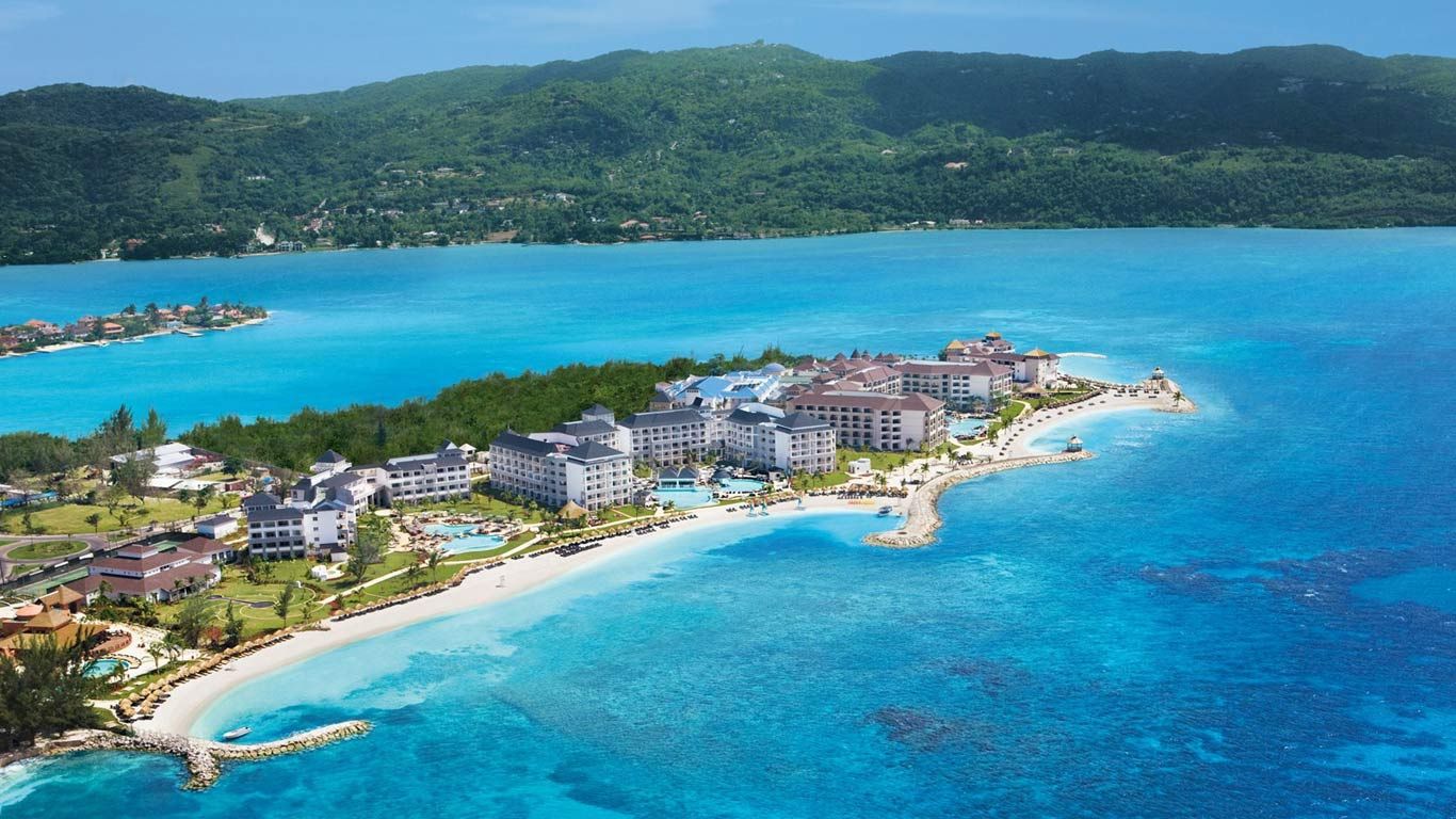 Aerial view of Secrets St. James resort in Montego Bay, Jamaica, showing beachfront buildings, curved white sand shoreline, and clear blue Caribbean waters.