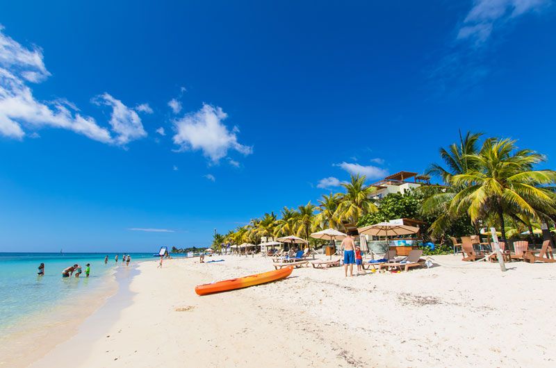 Sunny Roatán beach with white sand, calm turquoise water, palm trees, lounge chairs and umbrellas, people wading along the shoreline, and an orange kayak on the sand beneath a bright blue sky.