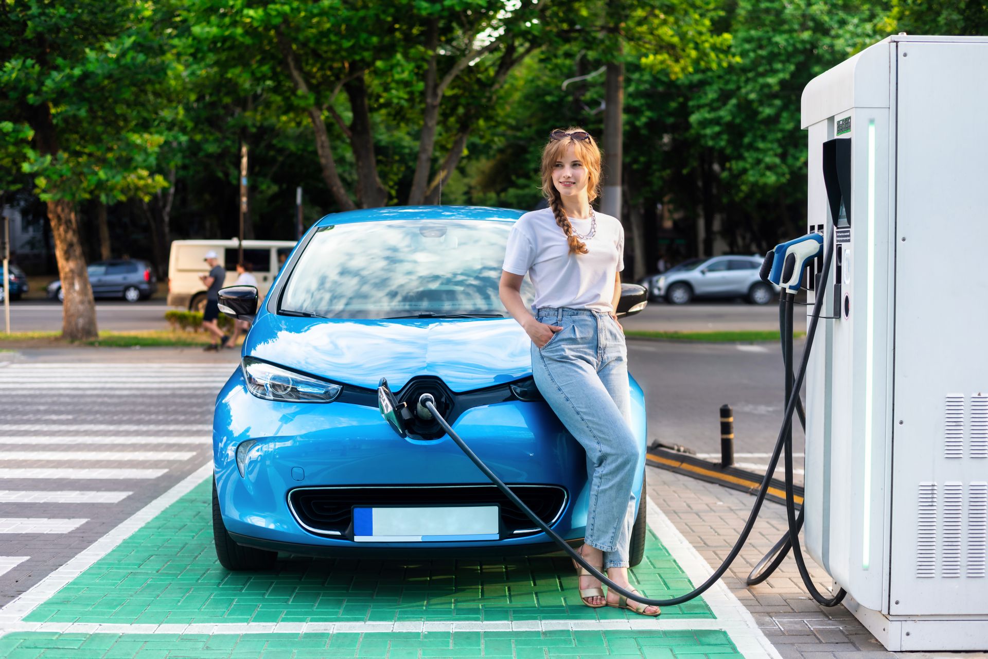 Jeune femme assise sur sa Renault Zoe qui est en train de recharger sur une borne de rechargement électrique publique Jeune femme assise sur sa Renault Zoe qui est en train de recharger sur une borne de rechargement électrique publique