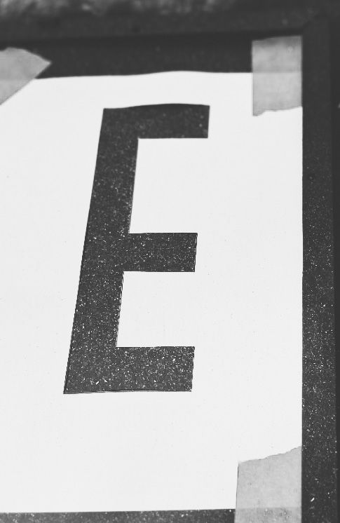 Large black letter “E” cut out as a stencil on white paper, taped down on a dark surface for a DIY garage sale sign project.