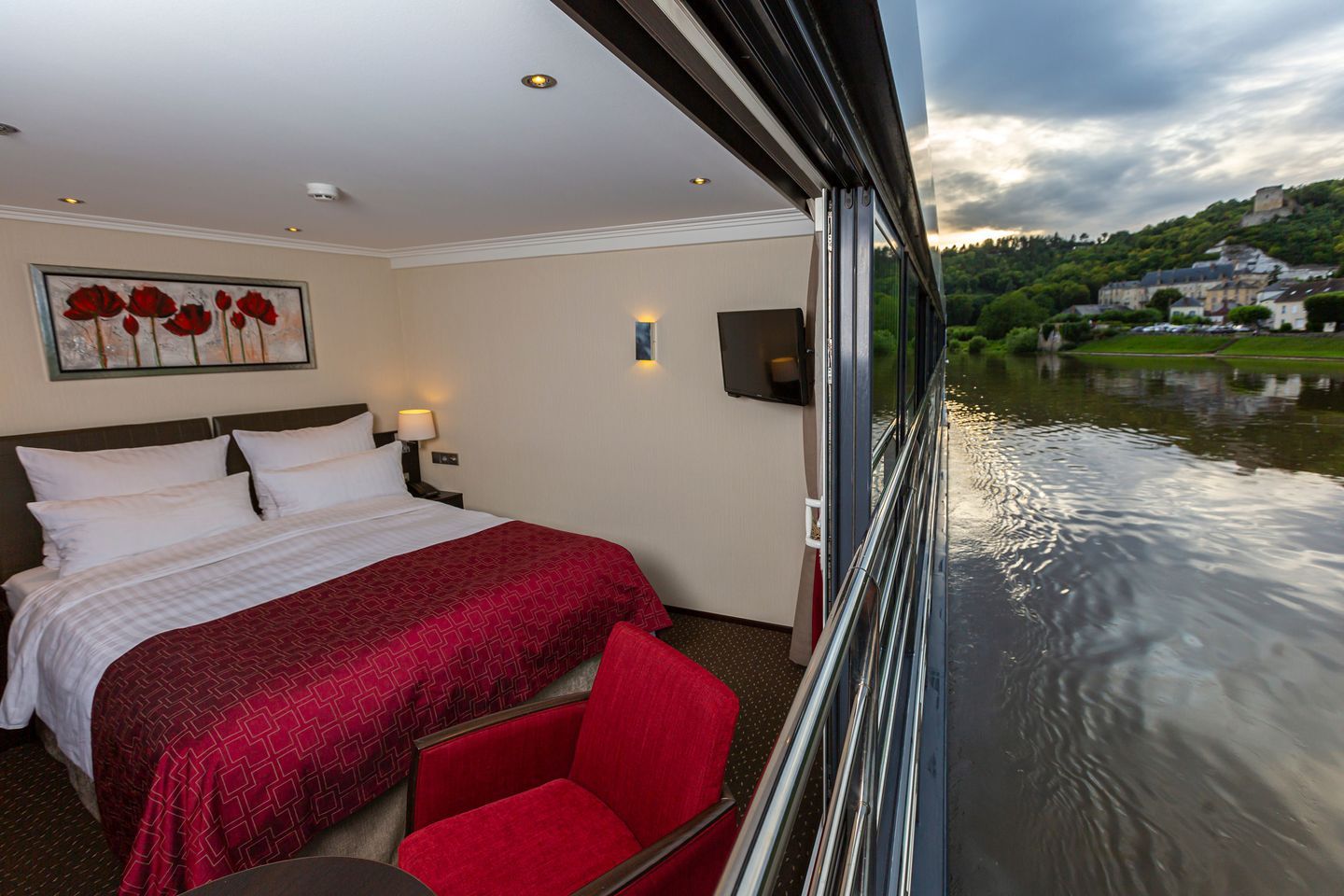 A cozy river cruise cabin with a large bed, red accents, and a window view overlooking a scenic riverbank with green hills and buildings at sunset. A cozy river cruise cabin with a large bed, red accents, and a window view overlooking a scenic riverbank with green hills and buildings at sunset.