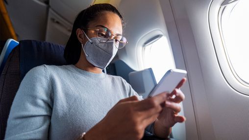 A traveler wearing a face mask and glasses sits on an airplane, looking at her smartphone — representing safe and mindful travel practices. A traveler wearing a face mask and glasses sits on an airplane, looking at her smartphone — representing safe and mindful travel practices.