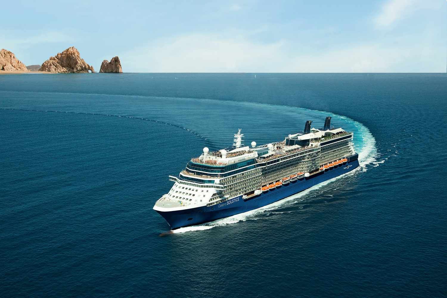 A Celebrity Cruises ship sails through crystal-clear blue waters near dramatic coastal rock formations on a sunny day, showcasing a luxury cruise vacation in a beautiful ocean setting.