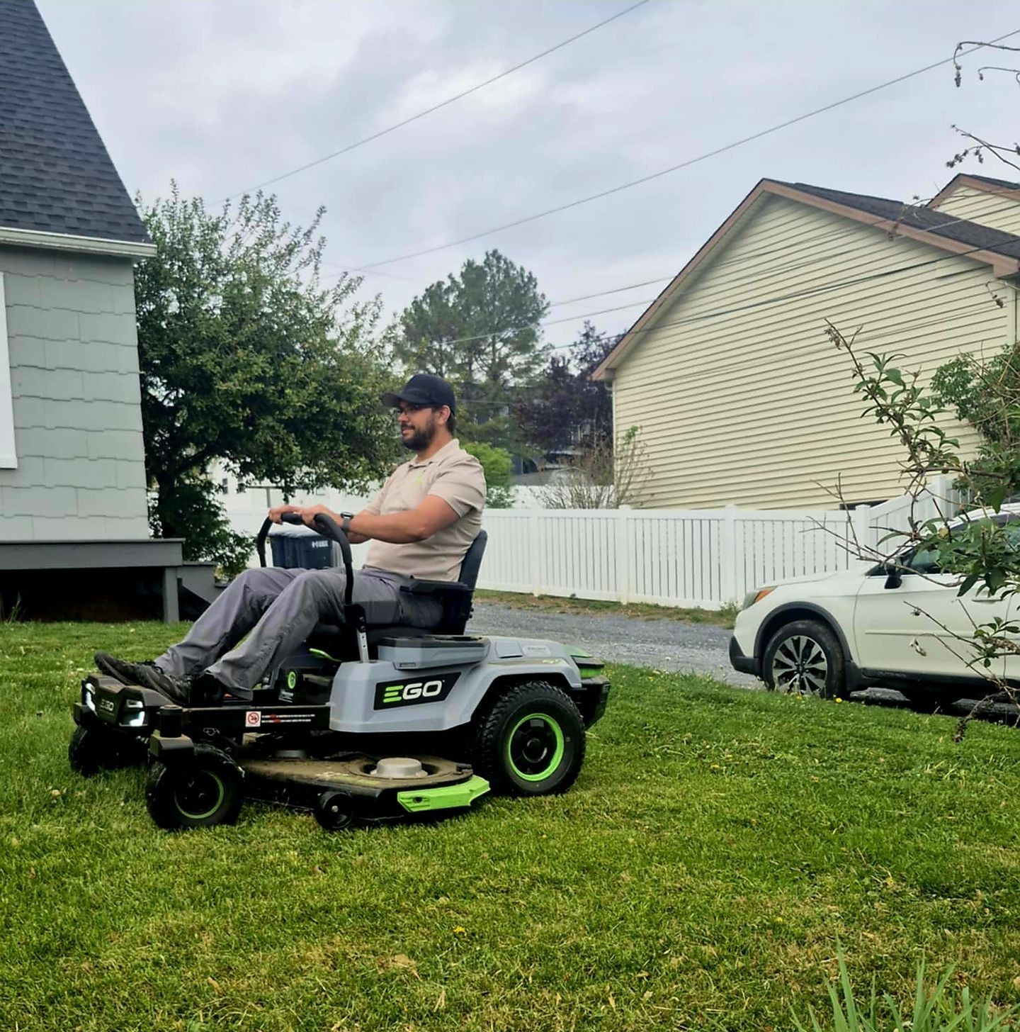 Person operating a riding lawn mower on a grassy lawn near a house and parked car.