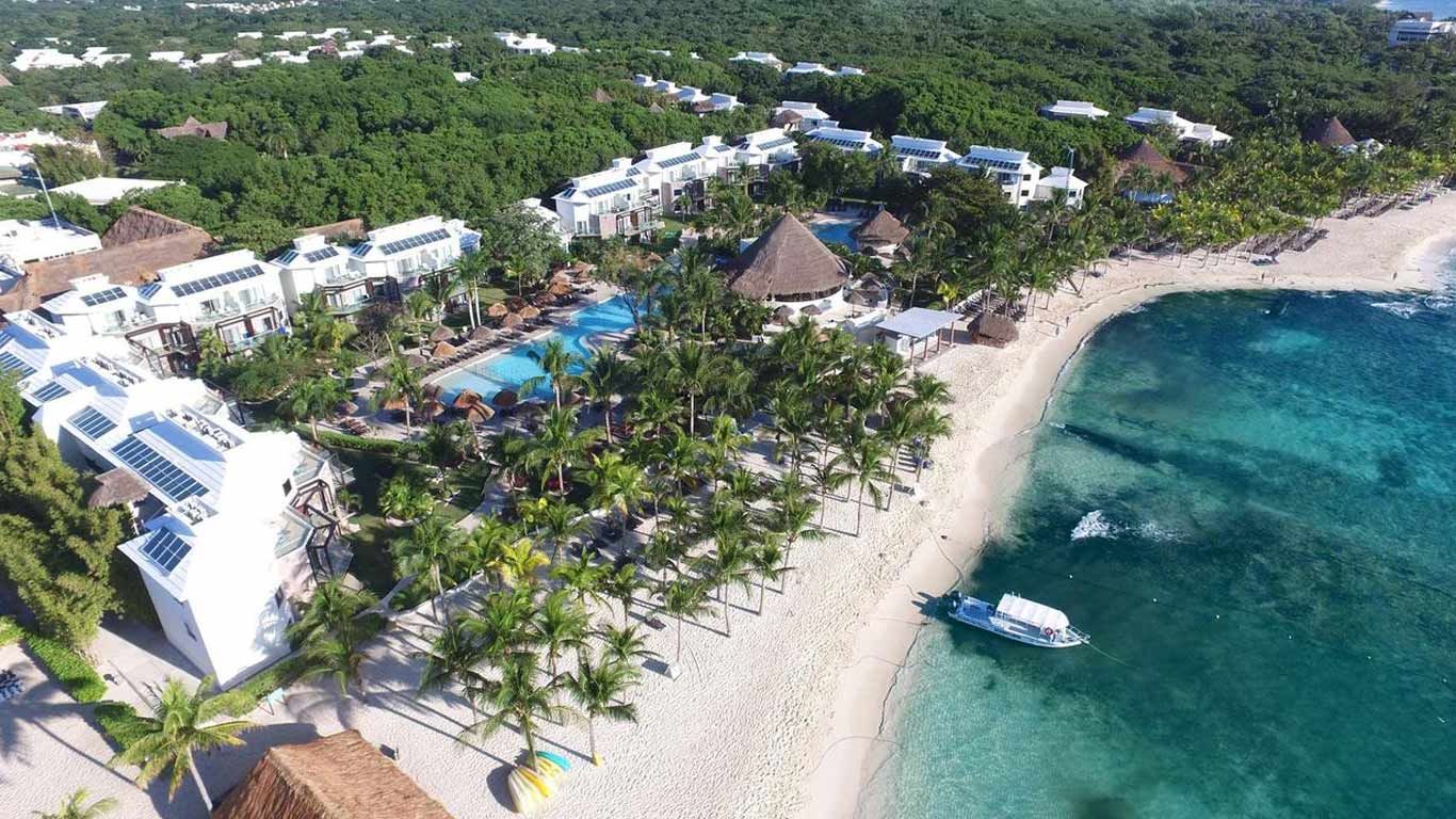 Aerial view of a tropical beachfront resort with white buildings, palm-lined sand, a central pool area, and clear turquoise ocean water along a curved shoreline, with dense green jungle stretching inland.