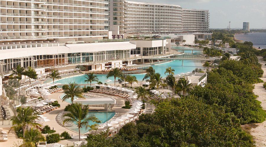 Elevated view of AVA Resort Cancun featuring multiple turquoise swimming pools, palm trees, white lounge chairs, and expansive oceanfront resort buildings along the Caribbean coastline.