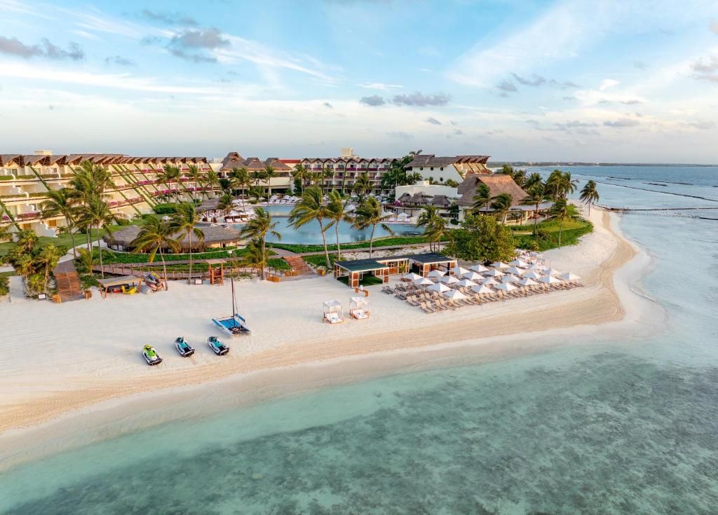 Aerial view of a beachfront resort with palm trees, a large lagoon-style pool, rows of lounge chairs and umbrellas on white sand, and calm turquoise water along the shoreline. Aerial view of a beachfront resort with palm trees, a large lagoon-style pool, rows of lounge chairs and umbrellas on white sand, and calm turquoise water along the shoreline.