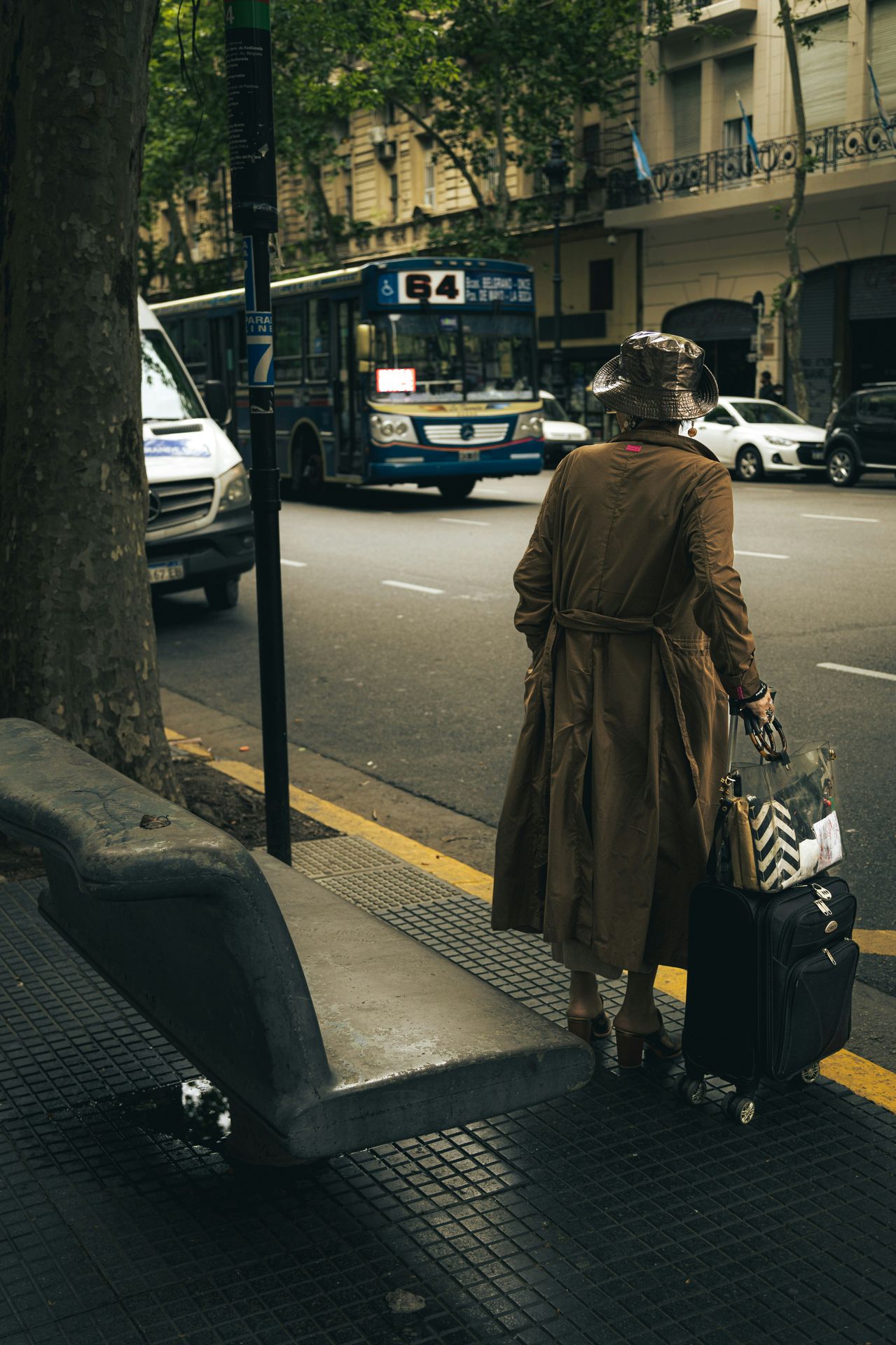 A woman in a trench coat and suitcase standing at a bus stop