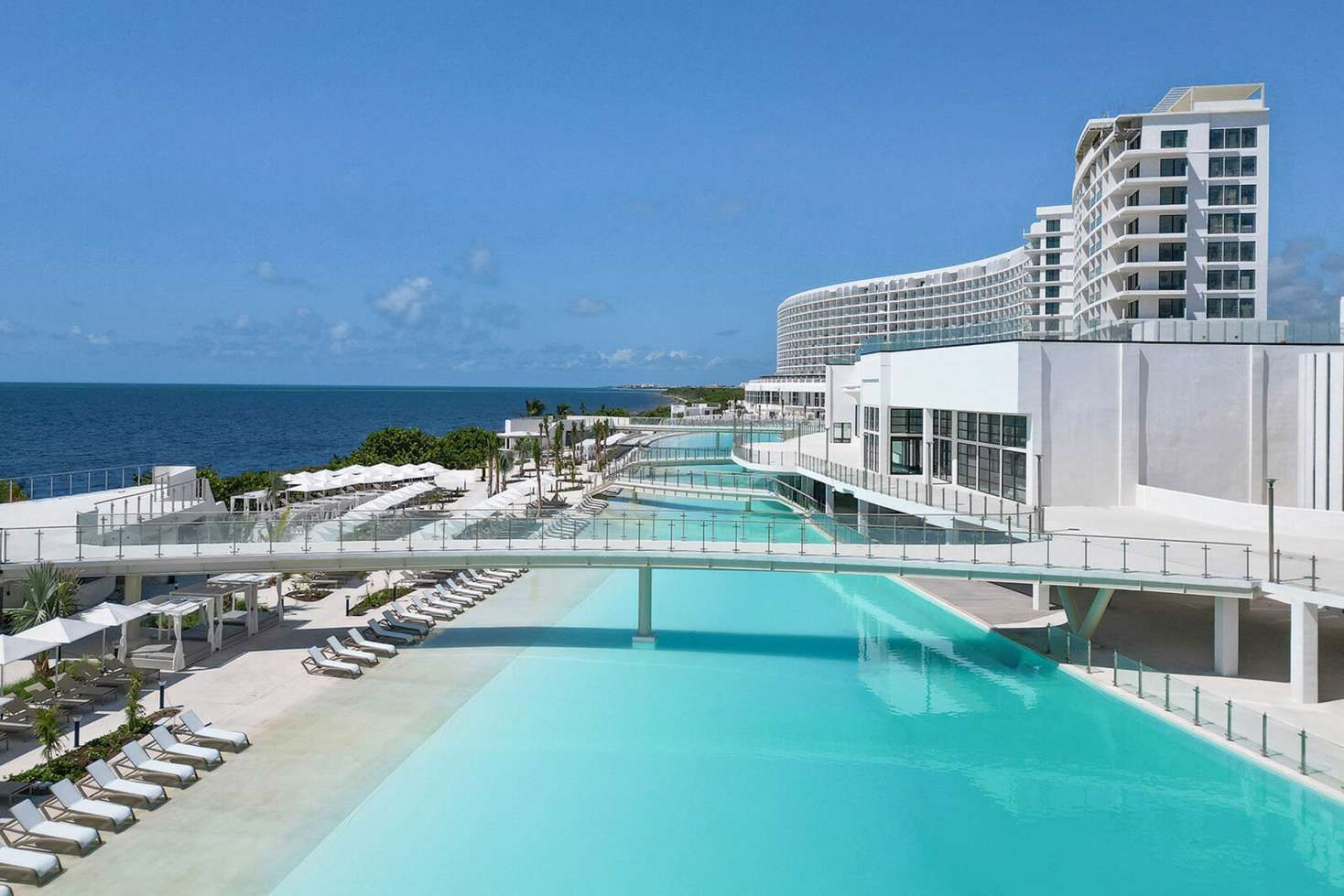Expansive turquoise swimming pools at AVA Resort Cancun, surrounded by lounge chairs, white umbrellas, and modern oceanfront buildings overlooking the Caribbean Sea.
