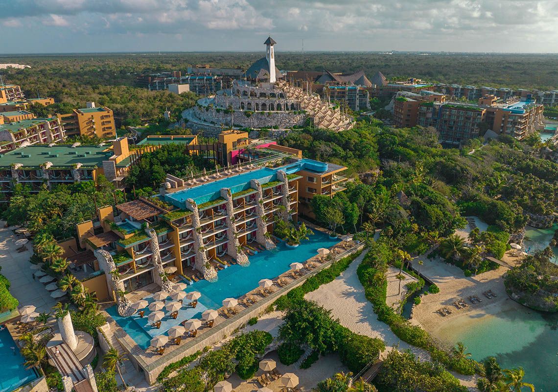 Aerial view of a sprawling Riviera Maya resort surrounded by lush jungle, featuring a multi-level turquoise pool, sandy beach coves, and a large stone, pyramid-like structure with a tower in the background under a partly cloudy sky.
