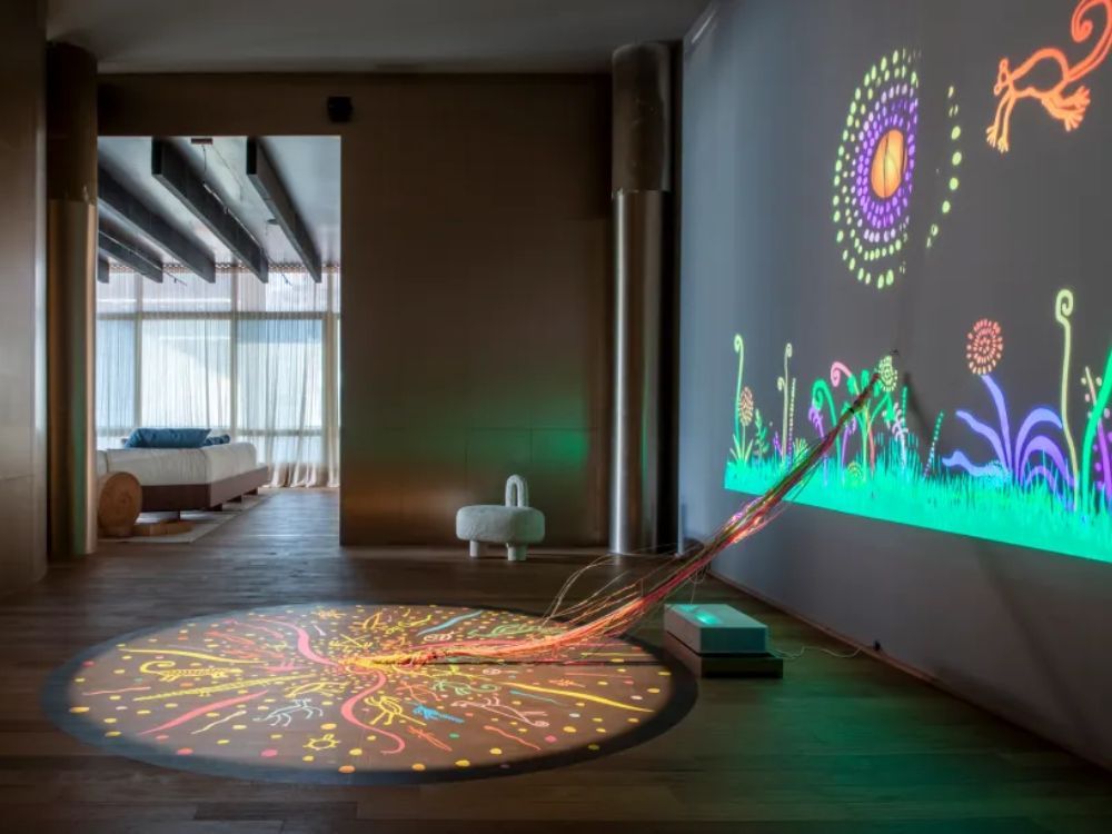 Sala escura com projeção de desenhos coloridos na parede e instalação de arte ligando a parede ao chão colorido