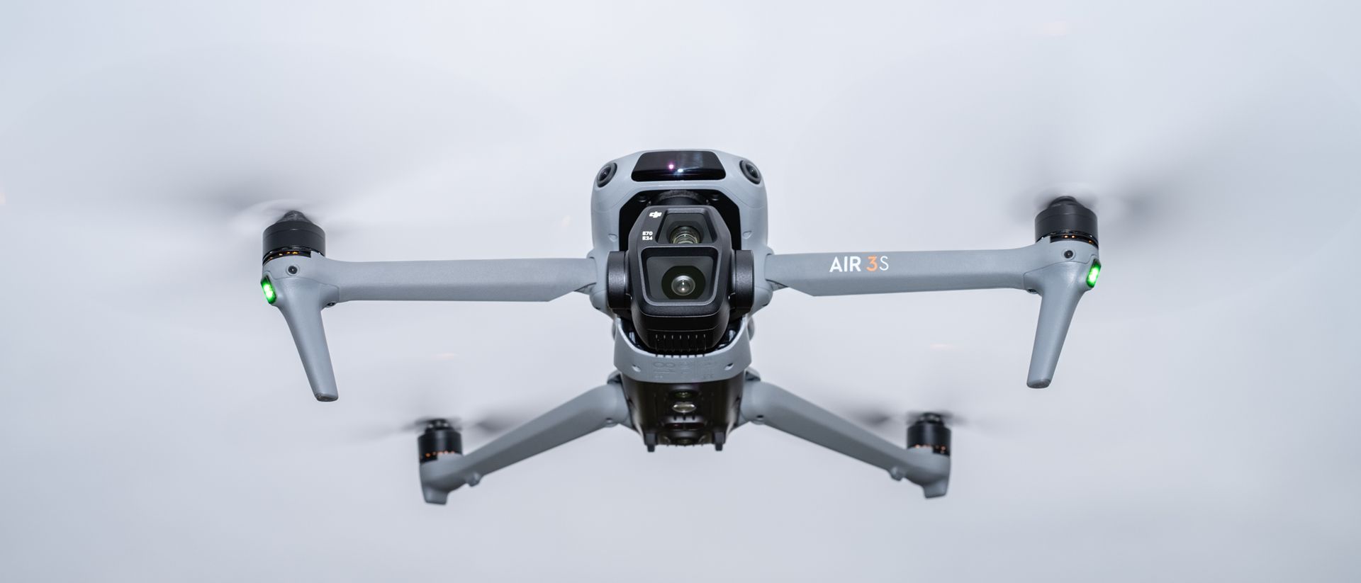 DJI Air 3S drone view showing gray body, dual-camera gimbal with 50MP wide-angle and medium telephoto lenses, black propellers with orange tips