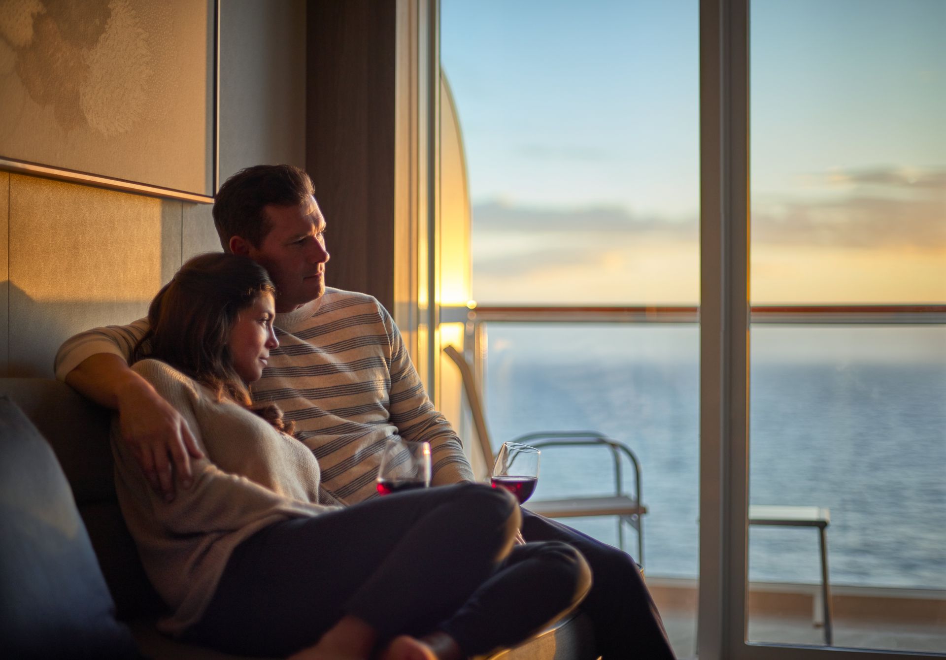 Couple relaxing on a stateroom sofa, enjoying wine and a golden sunset over the ocean through floor-to-ceiling windows.