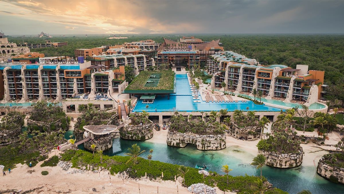 Aerial view of a modern Riviera Maya resort at sunset, surrounded by dense jungle, with a long central infinity pool, multiple terrace-style buildings, and a natural lagoon inlet with sandy beach and rocky edges in the foreground.