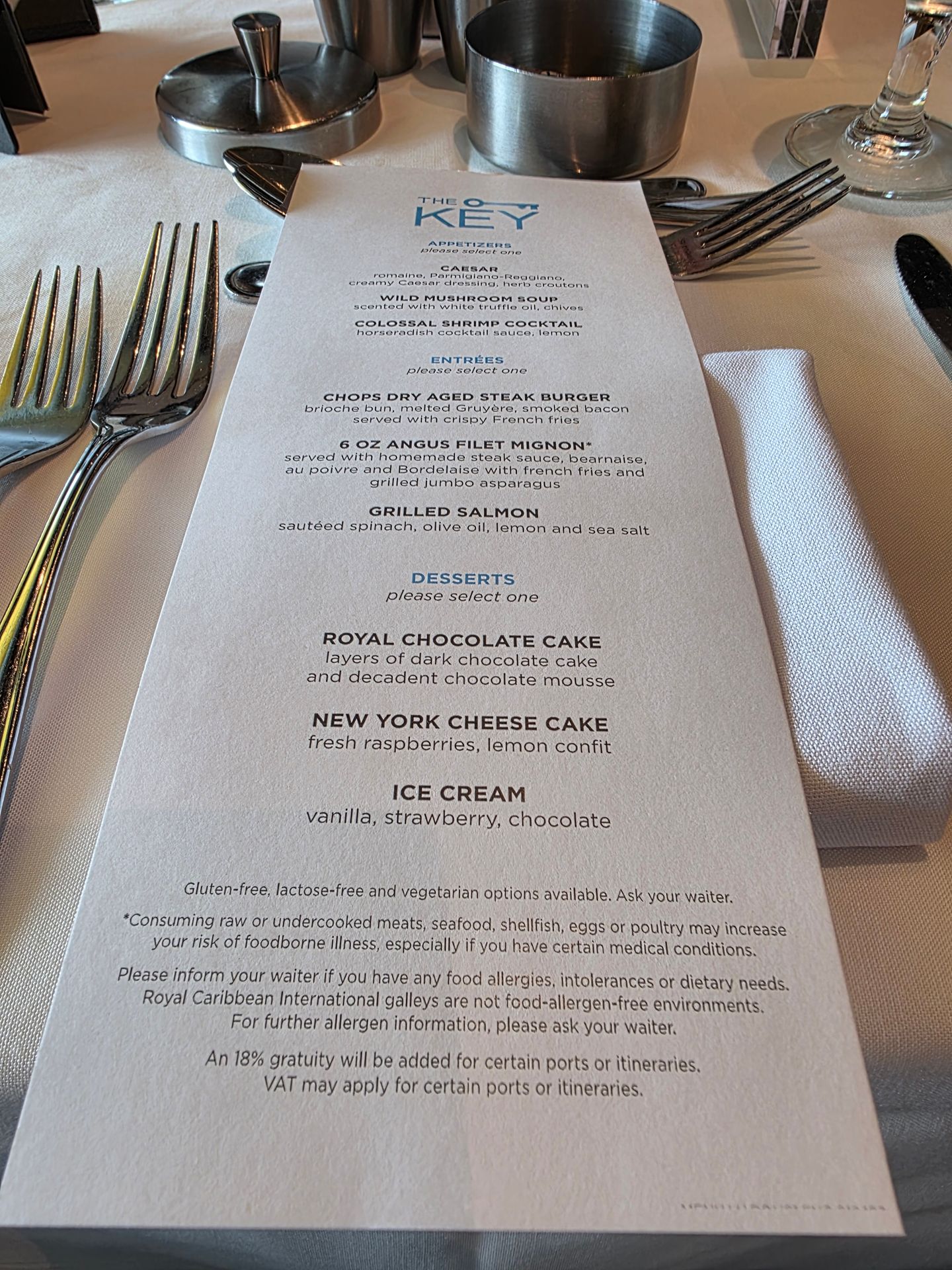 A close-up photo of a formal dining table with silverware and a printed menu from “The Key,” listing appetizers, entrées, and desserts on a white tablecloth. A close-up photo of a formal dining table with silverware and a printed menu from “The Key,” listing appetizers, entrées, and desserts on a white tablecloth.