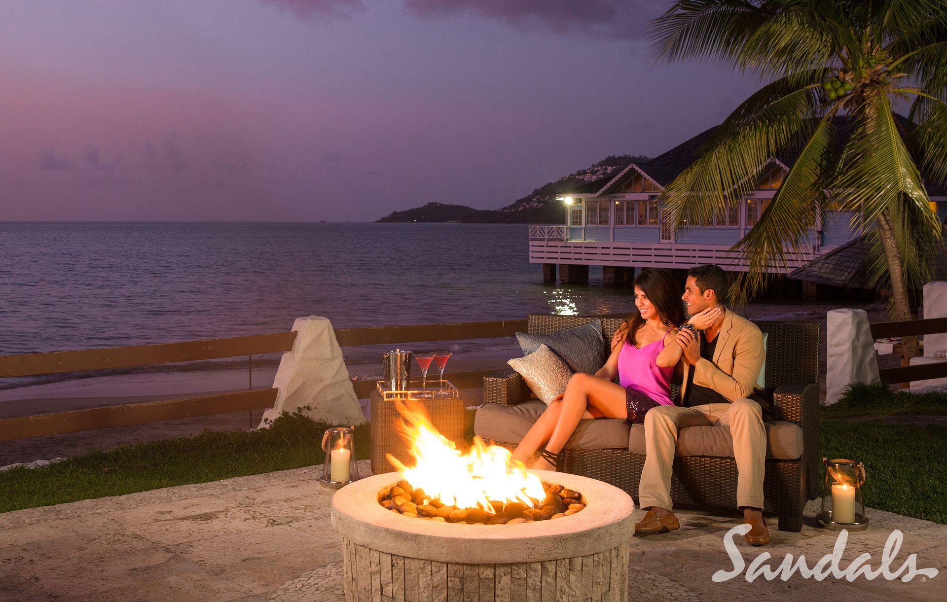 Couple relaxing by a fire pit on a beachfront patio at sunset, with cocktails on a side table and ocean views near overwater bungalows at a Sandals resort. Couple relaxing by a fire pit on a beachfront patio at sunset, with cocktails on a side table and ocean views near overwater bungalows at a Sandals resort.