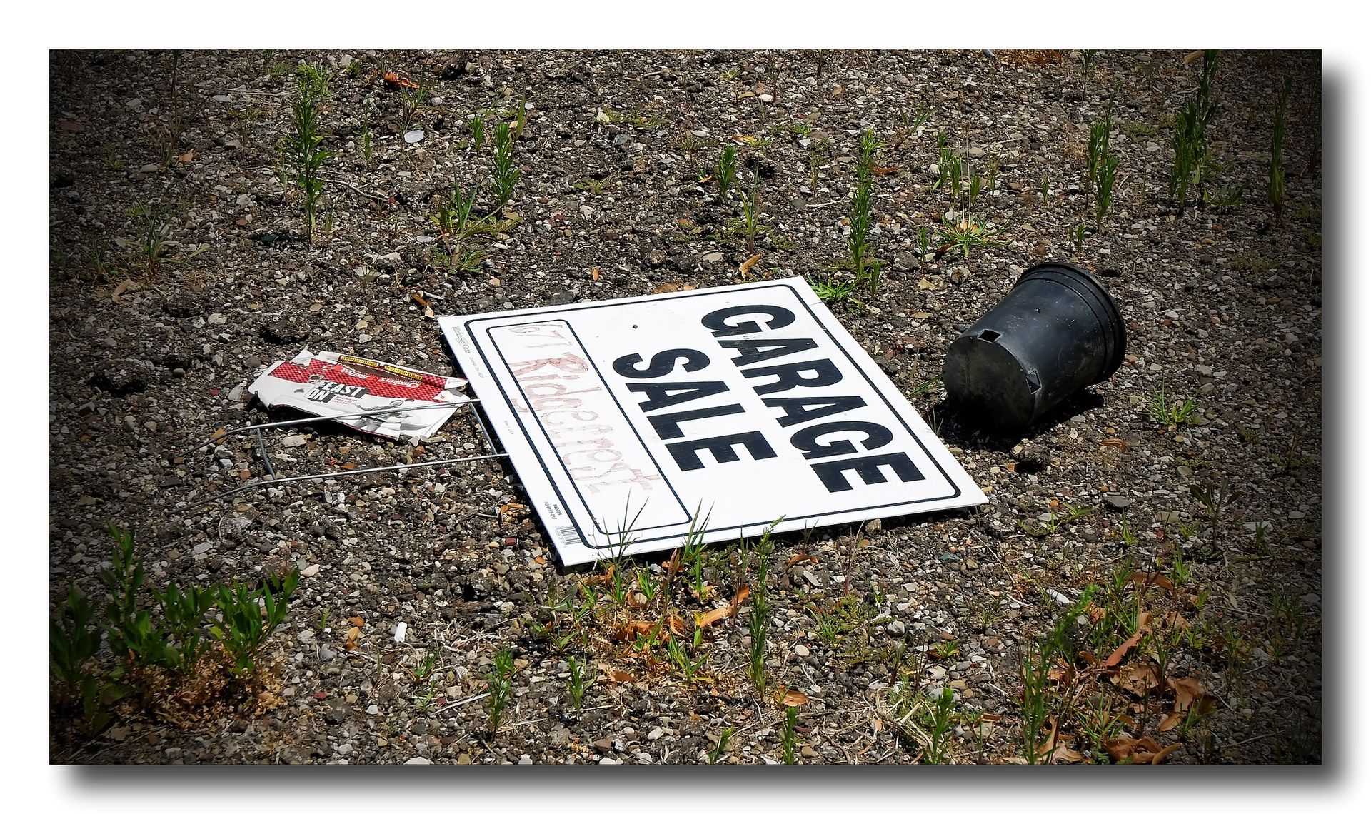 “Small black-and-white garage sale sign lying on the ground next to a broken plant pot and weeds”