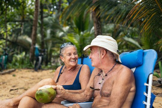 Older couple relaxing on tropical beach chairs, smiling at each other while holding a fresh coconut under palm trees.