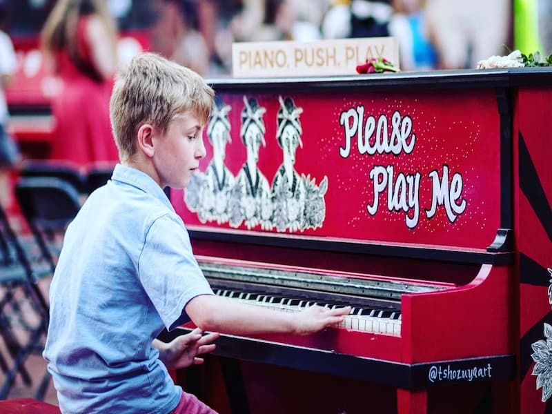 blonde boy in light blue collared shirt playing red piano that says 