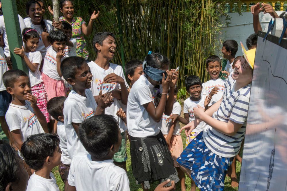 A joyful group of children laugh and cheer as they participate in a fun outdoor game together. One child is blindfolded while classmates and an adult encourage them, creating a moment filled with excitement, friendship, and shared laughter.
