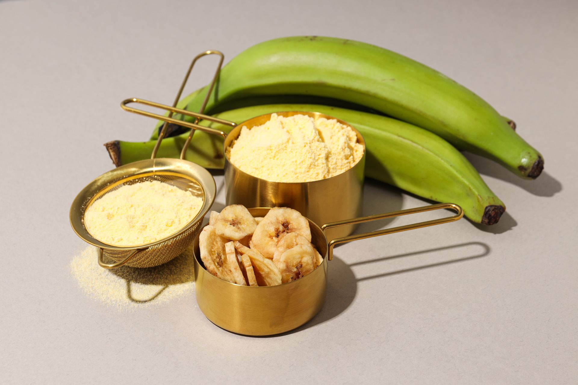 beurre de karité versé dans une passoire et un bol dorés, morceaux de banane dans un autre bol cuivré, et plusieurs bananes inclinées sur fond gris texturé