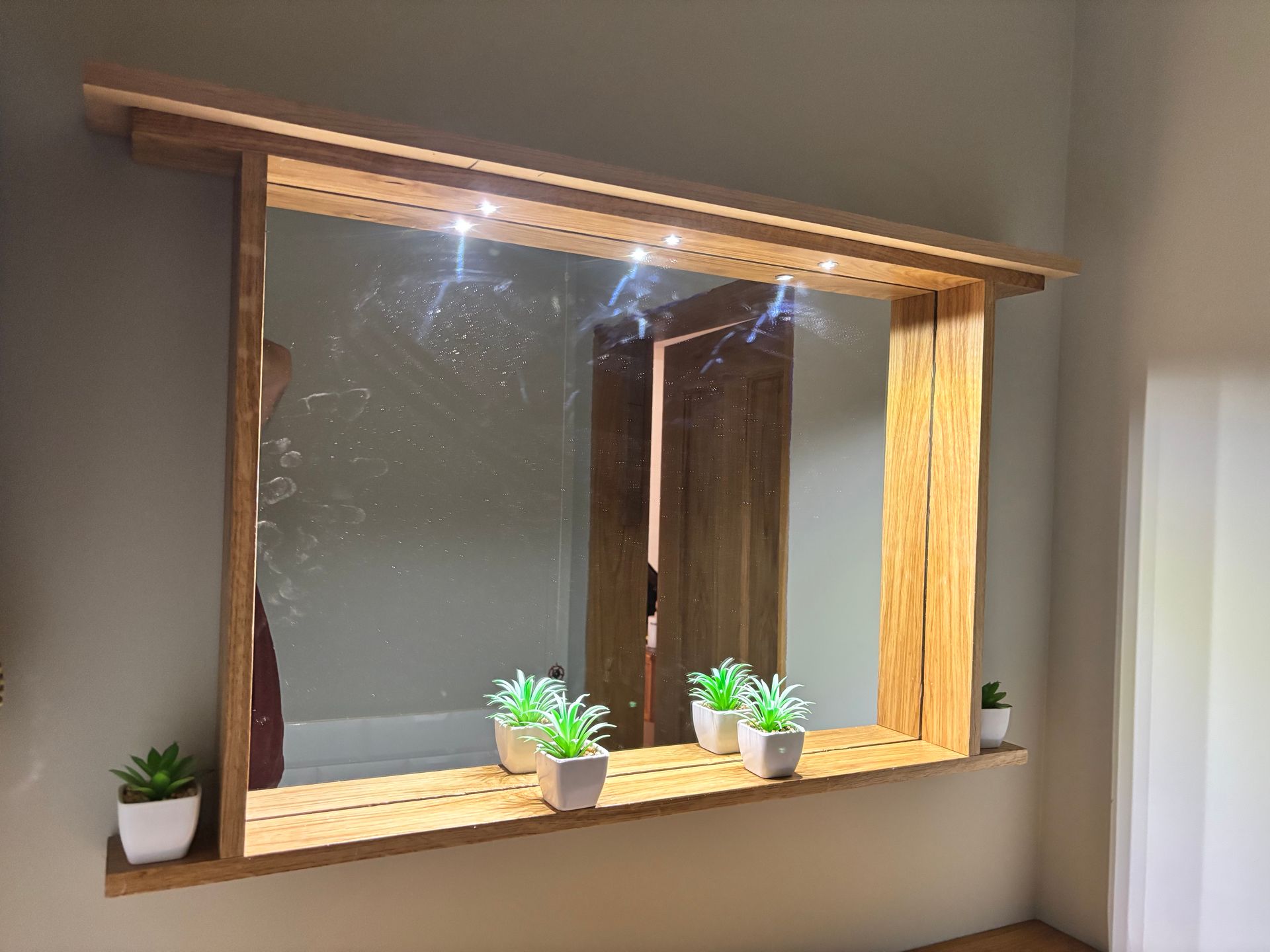 Finished oak mirror installed on the wall with integrated lighting and small potted plants on the shelf.