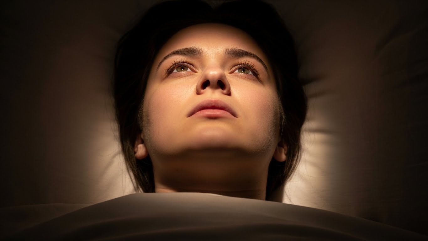 Mulher deitada olhando para cima no escuro, com expressão cansada e alerta, simbolizando insônia, esgotamento emocional e mente que não desacelera. Mulher deitada olhando para cima no escuro, com expressão cansada e alerta, simbolizando insônia, esgotamento emocional e mente que não desacelera.