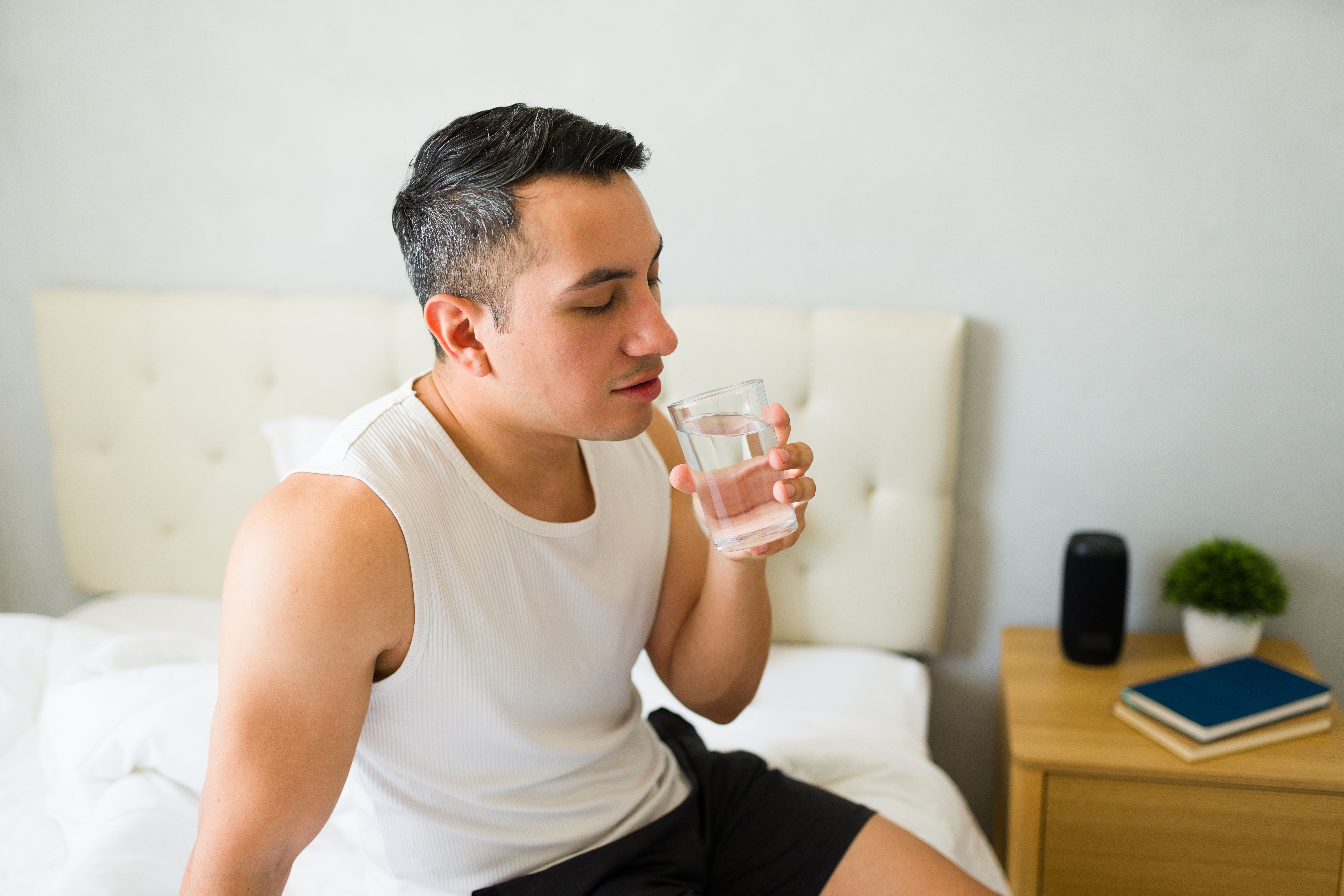 Young man is sitting on his bed in his bedroom, drinking a glass of water after waking up in the morning