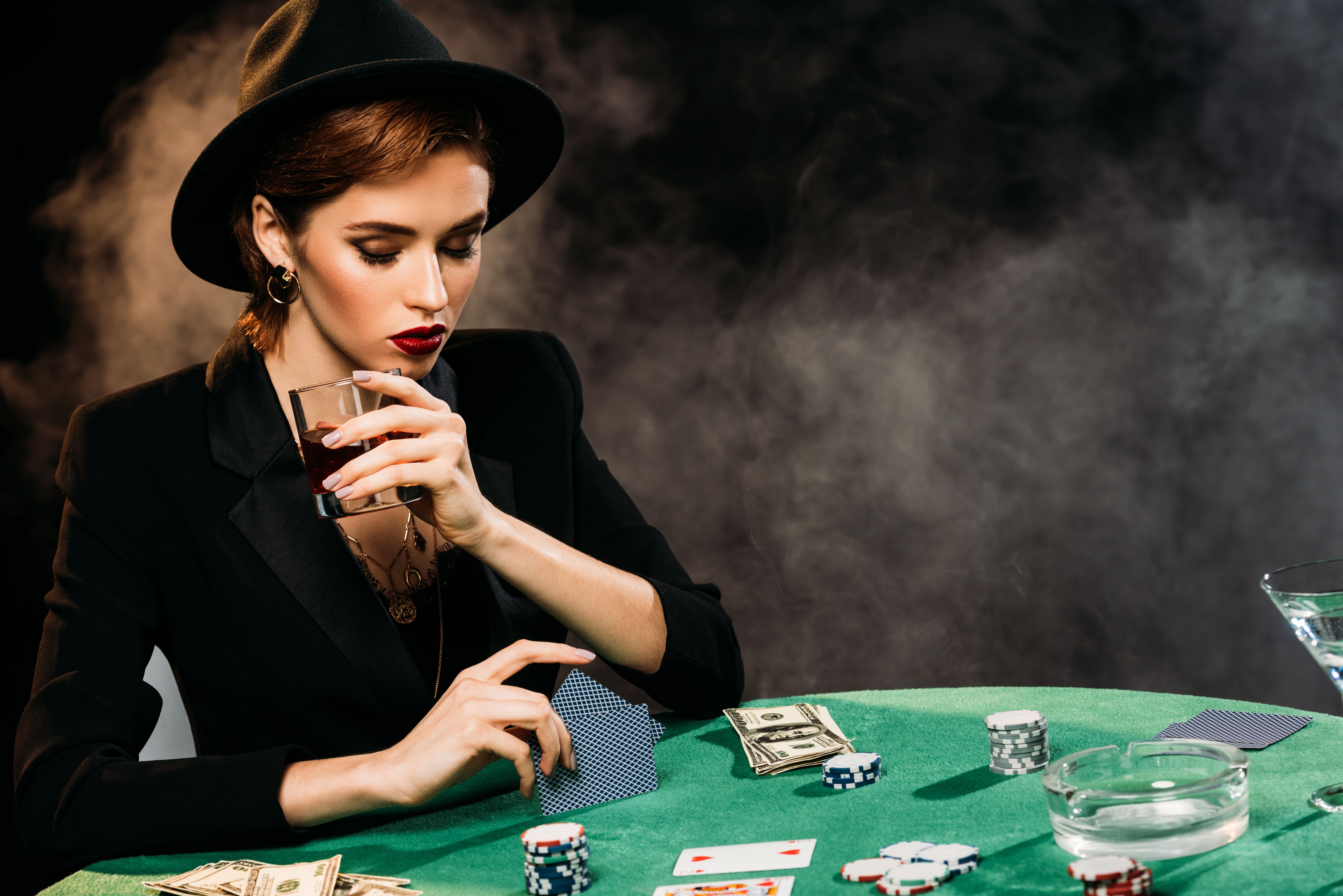 pensive attractive girl in jacket and hat playing poker and holding glass of whiskey in casino